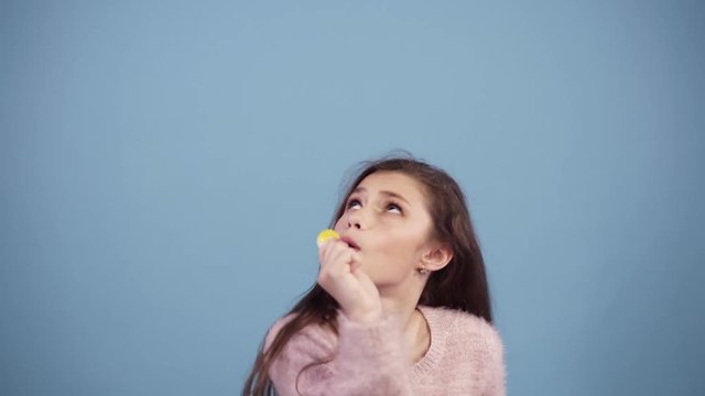 Funny girl in pink sweater having fun, dancing, jumping and singing to yellow lollipop. Portrait of a pretty young girl singing to lollipop over blue background. The girl sings in a candy, like a