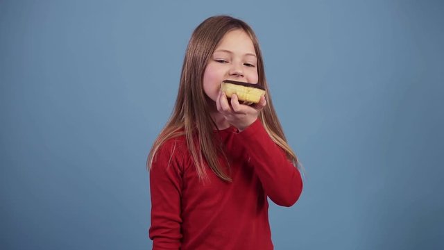 Little girl in red sweater is eating a chocolate donut on a colored background. Enjoying the taste. Child 7-10 years old, fair hair. Shooting in the studio. Food concept