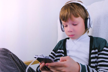 Boy is watching a video in headphones in mobile phone sitting in armchair