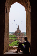 A young man sitting in a temple at the lovely sunrise with balloons on top of the temples in Bagan,...