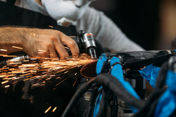 Worker cutting, grinding and polishing motorcycle metal part with sparks indoor workshop, close-up.