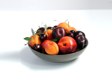 Fruit bowl. Seasonal fruits with green leaves in a bowl over white background, top view. Organic fresh fruit, peach, plum, aprocot, cherry.