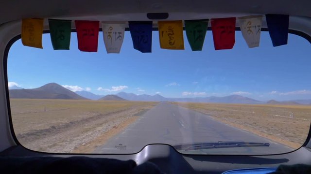 SLOW MOTION, CLOSE UP: Spectacular view of an empty road in Tibet through the rear window of a car. Colorful prayer flags hang above a car window offering a stunning view of the Himalayan Plateau.