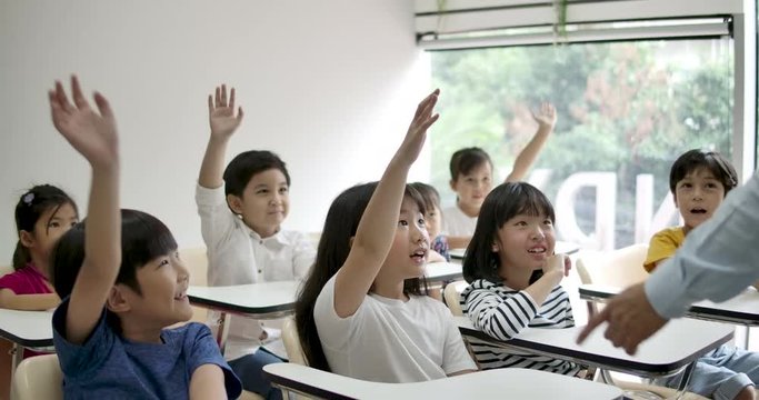 Group of joyful asian elementary students raising their arms to answer a question from teacher at classroom. Kindergarten pre school concept.