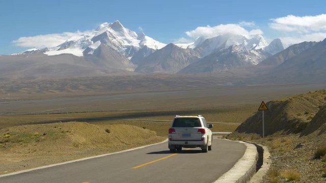 White SUV drives down an empty asphalt road leading to the snowy mountains in Tibet. Locals driving across the breathtaking plains of the Himalayan Plateau. Travelers exploring the Tibetan fields
