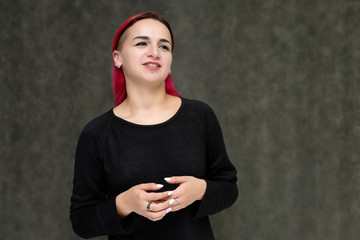 Portrait to the waist of a pretty girl with red hair on a gray background in a black jacket. Standing right in front of the camera in a studio with emotions, talking, showing hands, smiling