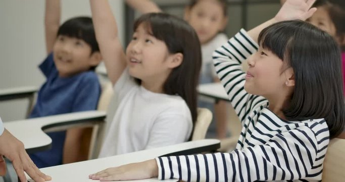 Group of joyful asian elementary students raising their arms to answer a question from teacher at classroom. Kindergarten pre school concept.