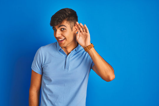 Young indian man wearing casual polo standing over isolated blue background smiling with hand over ear listening an hearing to rumor or gossip. Deafness concept.