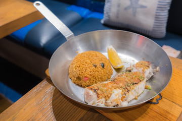 Grilled fish steak served with fried rice and lemon in a deep pan placed on a wooden board.