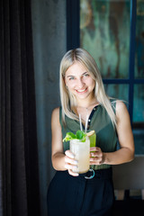 A young beautiful blonde girl stands in the middle of a cozy cafe and drinks a cooing drink. Rest in the summer heat