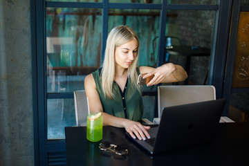 A young beautiful blonde girl sits in a cozy cafe and works for a laptop, drinks a cooling drink.