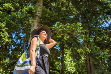 Fototapeta premium Attractive happy young woman with hat and bag enjoying her time outside in nature near trees on sunny day