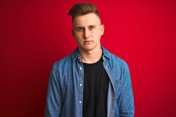 Young handsome man wearing denim shirt standing over isolated red background Relaxed with serious expression on face. Simple and natural looking at the camera.