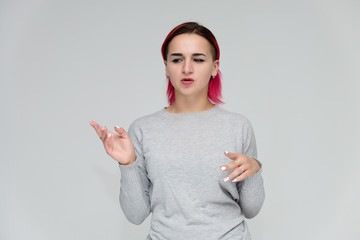 Portrait to the waist of a pretty girl with red hair on a white background in a gray sweater. Standing right in front of the camera in a studio with emotions, talking, showing hands, smiling