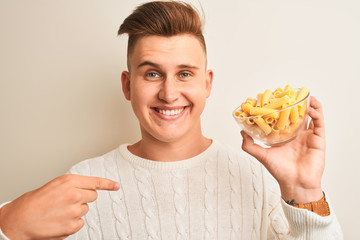 Young handsome man holding bowl with dry pasta standing over isolated white background with surprise face pointing finger to himself