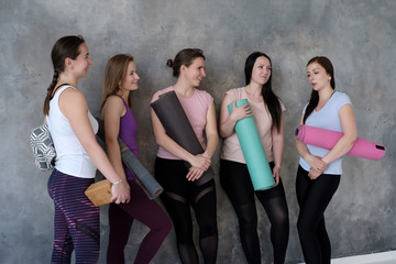 Caucasian women stand near wall with rubber mats in hands, have fun waiting for yoga class. Having chat before lesson or workout in fitness studio together