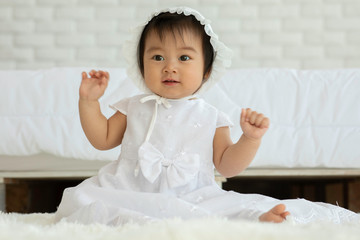 Fototapeta premium Cute Sweet Adorable Asian Baby wearing white dress sitting on Carpet smiling and playing with happiness emotional in cozy bedroom,Healthy Baby Concept