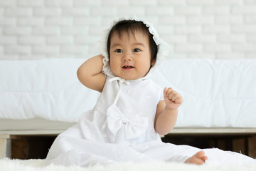 Fototapeta premium Cute Sweet Adorable Asian Baby wearing white dress sitting on Carpet smiling and playing with happiness emotional in cozy bedroom,Healthy Baby Concept
