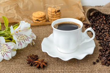 White peruvian lily flower, hot coffee in white elegant cup with saucer, sweet cookies and scattering of coffee beans on rustic sackcloth a background. Morning hot beverage.