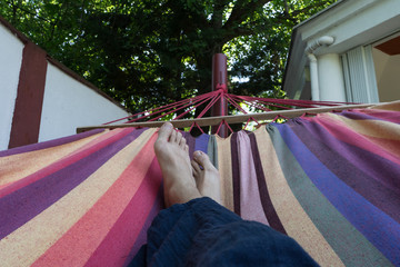 Guy lying down in a colorful Hammock resting barefoot.Crossed legs of a man taking nap in blue hippie pants in a summer garden of a house with a big tree. nude feet close up