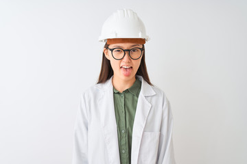 Young chinese engineer woman wearing coat helmet glasses over isolated white background sticking tongue out happy with funny expression. Emotion concept.