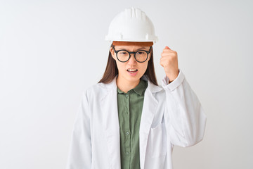 Young chinese engineer woman wearing coat helmet glasses over isolated white background angry and mad raising fist frustrated and furious while shouting with anger. Rage and aggressive concept.