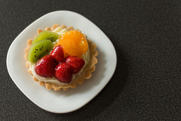 Cake with cream, raspberries, kiwi and orange on a black background. Pastry with fruits on top. Sweet dessert. Copy space.