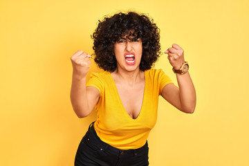 Young arab woman with curly hair wearing t-shirt standing over isolated yellow background angry and mad raising fists frustrated and furious while shouting with anger. Rage and aggressive concept.