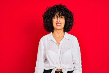 Young arab woman with curly hair wearing white casual shirt over isolated red background winking...