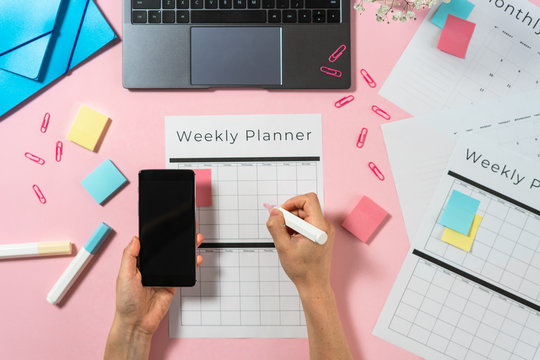 Woman holding smartphone and writing at planner on pink pastel background  with laptop and stationery