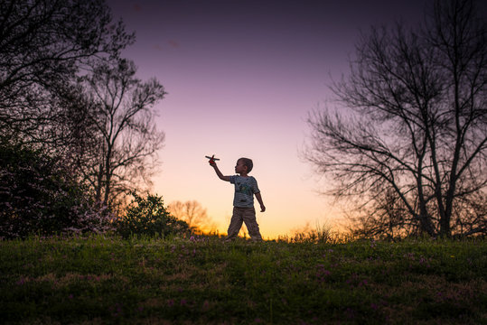 Little boy silohette playing w/ an airplane and sunset behind w/ trees