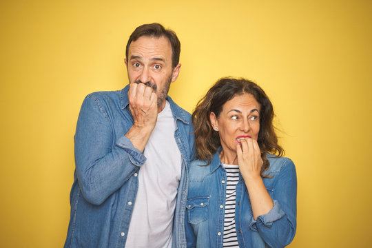 Beautiful middle age couple together wearing denim shirt over isolated yellow background looking stressed and nervous with hands on mouth biting nails. Anxiety problem.