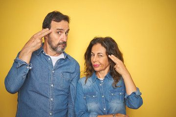 Obraz premium Beautiful middle age couple together standing over isolated yellow background Shooting and killing oneself pointing hand and fingers to head like gun, suicide gesture.