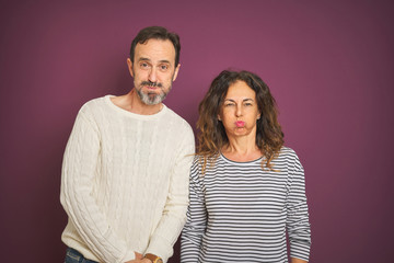 Beautiful middle age couple wearing winter sweater over isolated purple background puffing cheeks with funny face. Mouth inflated with air, crazy expression.