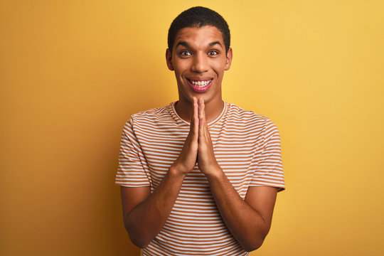 Young handsome arab man wearing striped t-shirt standing over isolated yellow background praying with hands together asking for forgiveness smiling confident.