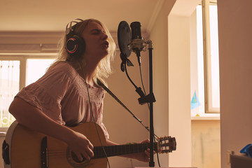 Beautiful blonde singer girl in headphones with a guitar in home recording studio sings a song into a microphone