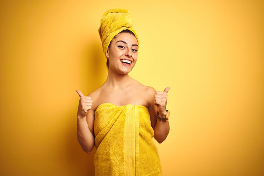 Young beautiful woman wearing towel after shower over isolated yellow background success sign doing positive gesture with hand, thumbs up smiling and happy. Cheerful expression and winner gesture.