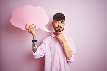 Young man with tattoo holding cloud speech bubble standing over isolated pink background serious...