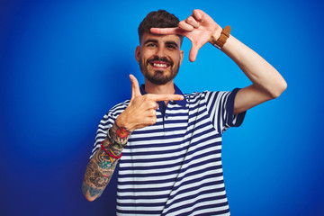 Young man with tattoo wearing striped polo standing over isolated blue background smiling making frame with hands and fingers with happy face. Creativity and photography concept.