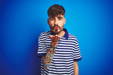 Young man with tattoo wearing striped polo standing over isolated blue background looking at the camera blowing a kiss with hand on air being lovely and sexy. Love expression.