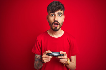 Young gamer man with tattoo playing video game standing over isolated red background scared in...