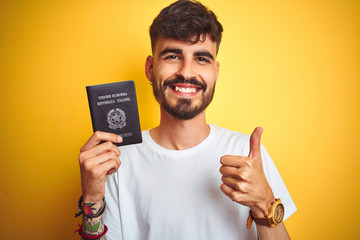 Young man with tattoo wearing Italy Italian passport over isolated yellow background happy with big smile doing ok sign, thumb up with fingers, excellent sign