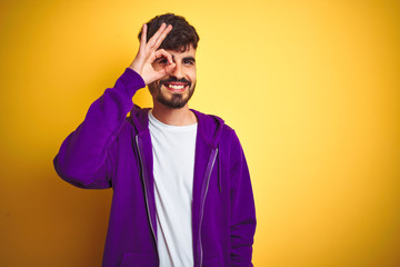 Obraz premium Young man with tattoo wearing sport purple sweatshirt over isolated yellow background doing ok gesture with hand smiling, eye looking through fingers with happy face.