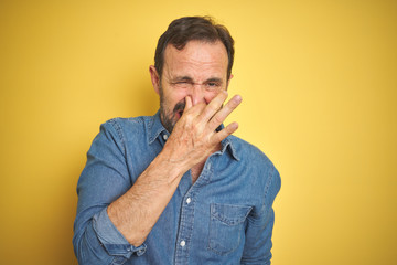 Handsome middle age senior man with grey hair over isolated yellow background smelling something...