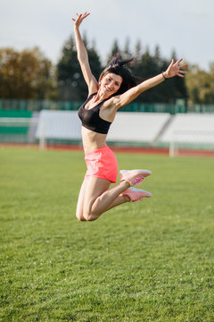 Attractive sporty happy brunette woman in pink shorts and top makes a high jump in sun rays at the stadium looking at the camera and smiling