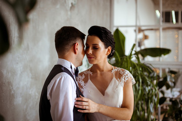 Beautiful bride in an elegant dress the groom in a waistcoat in a stylish interior with tropical plants against a concrete wall