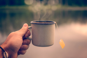 Male hand holding a mug of tea on the background of the lake