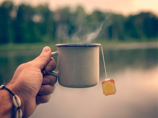 Male hand holding a mug of tea on the background of the lake