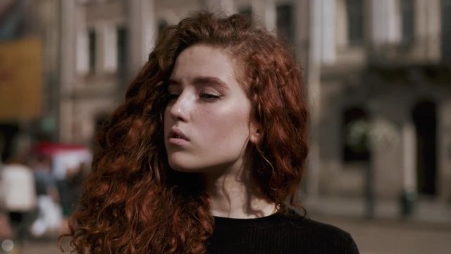Serious young woman with red curly hair look at camera stand walking in the city streets portrait attractive slow motion summer face sunset beautiful lady outdoor close up slow motion