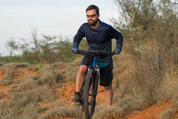 Fototapeta premium Cyclist in shorts and jersey on a modern carbon hardtail bike with an air suspension fork rides off-road on the orange-red hills at sunset evening in summer 